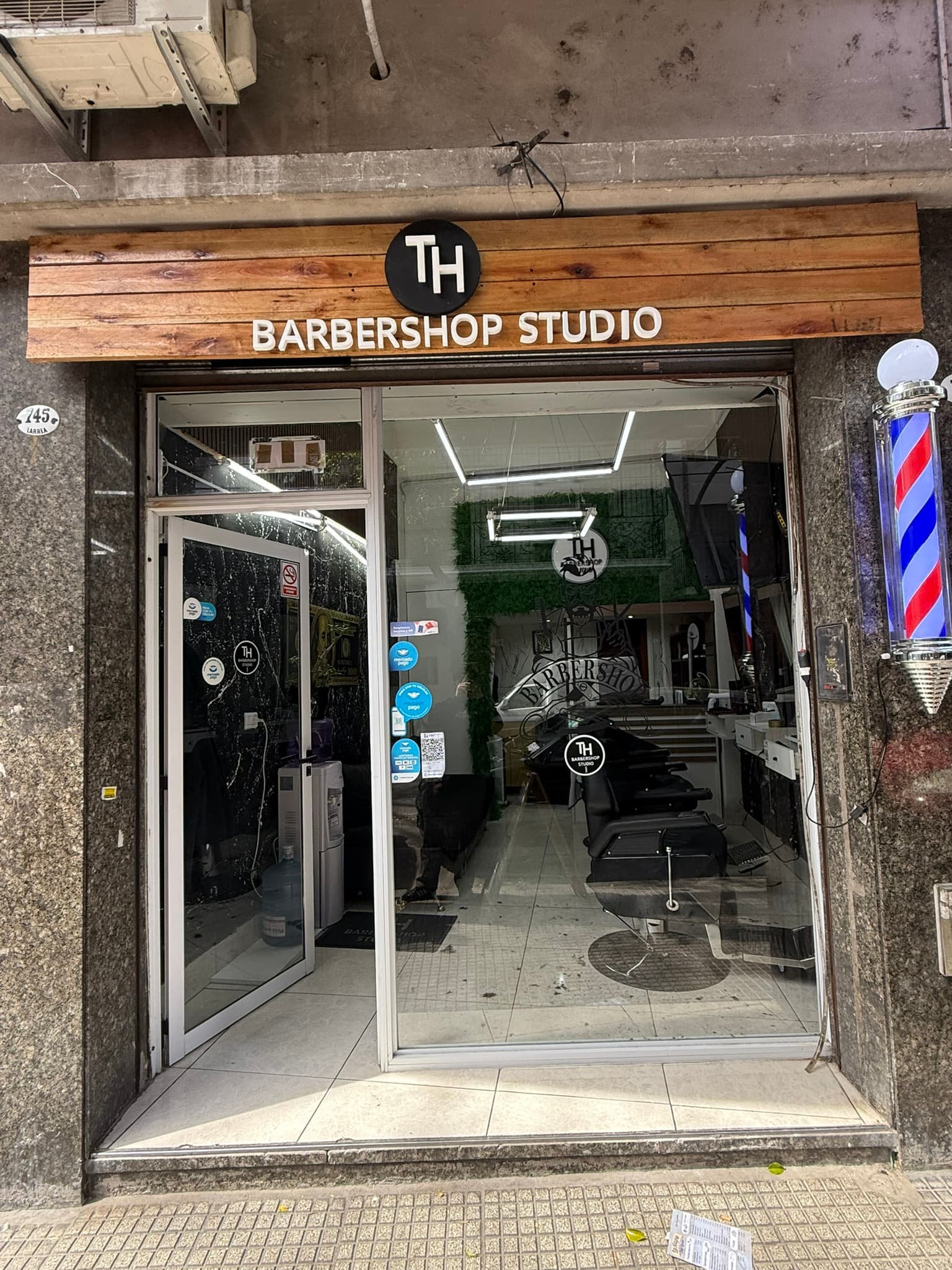 Fachada de TH Barbershop Studio: cartel de madera con el logo, poste de barbero y el local visto a través de la vidriera